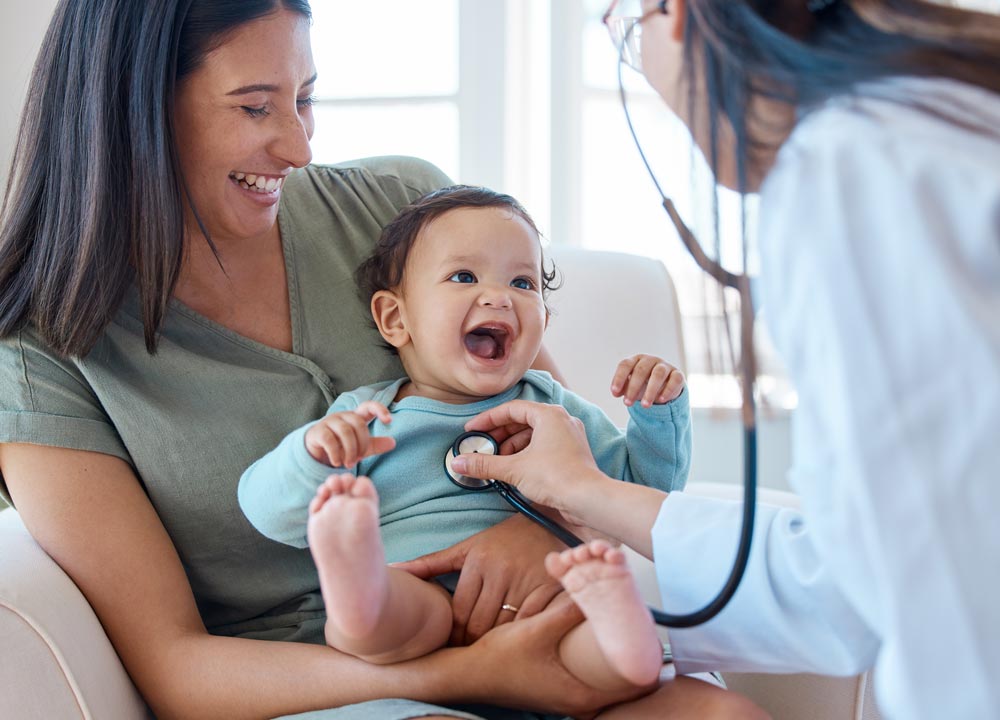 A mother and her baby smiling during a pediatric check-up, as a doctor examines the infant with a stethoscope.