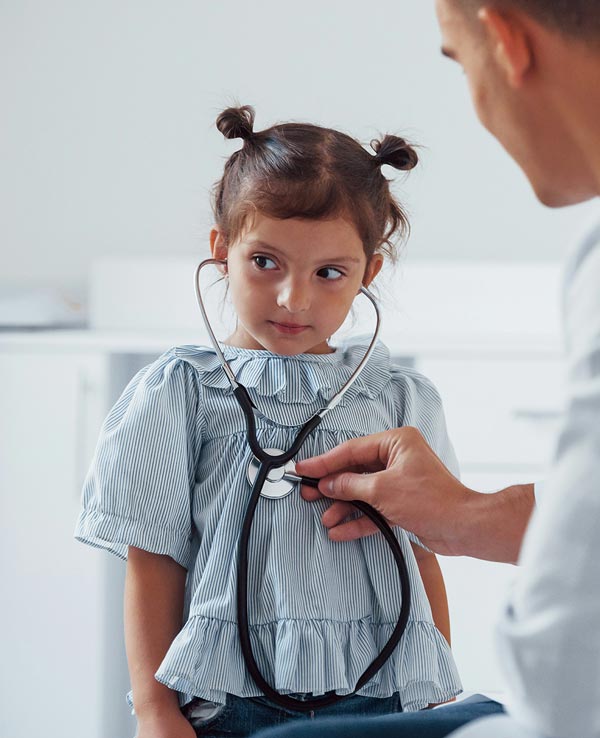 A young girl with pigtails looks curiously at a doctor using a stethoscope in a bright, modern medical setting.