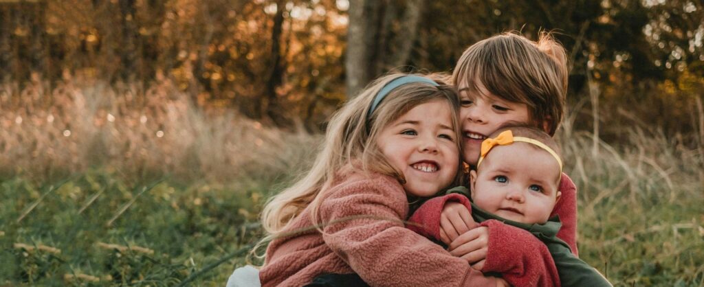 Three young children embrace each other in a grassy field, smiling and enjoying a warm, autumn day.