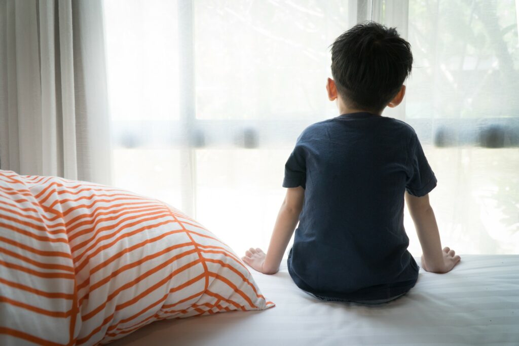 Child sits on a bed, facing away and looking out the window.