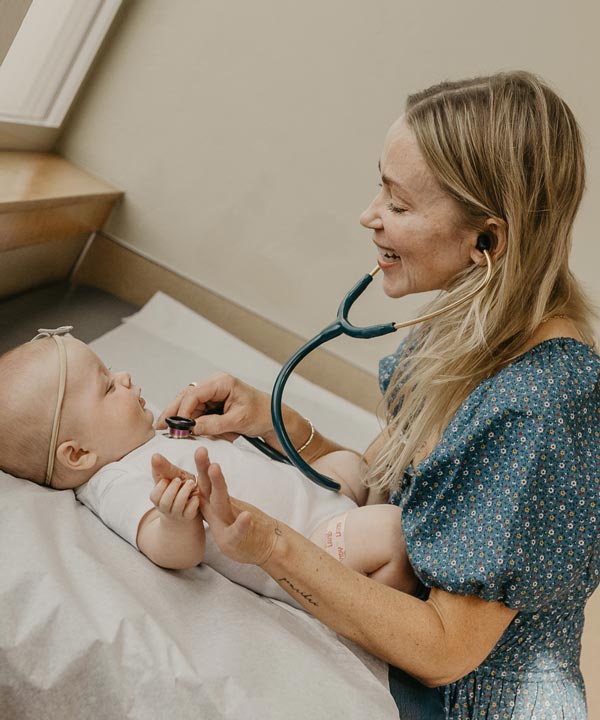 A woman joyfully examines a baby using a stethoscope in a bright medical setting.