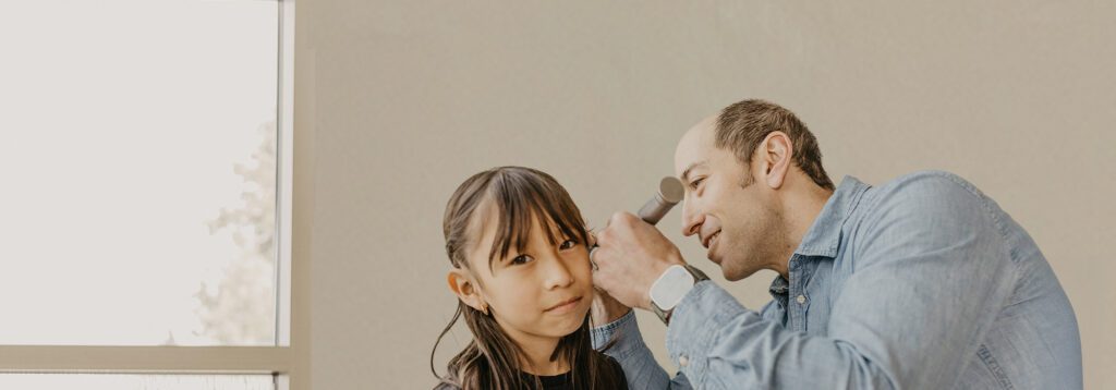 A man applies makeup to a young girl, both appearing to enjoy a lighthearted moment in a bright, airy room.