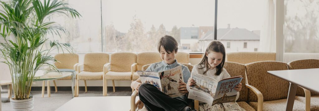Two individuals sit in a cozy waiting area, happily reading magazines, with a large window and a plant nearby.